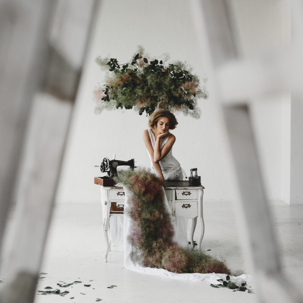 Woman in white clothes sits in a room with flowers and sewing machine